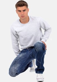 Felpa grigia abbinata a jeans denim blu con dettagli di cucitura, vestibilità ampia e finitura sbiadita. Sneakers bianche completano l'outfit.