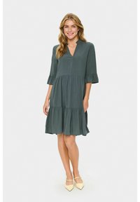 Rochie verde lungime până la genunchi, cu decolteu în V, mâneci trei sferturi și accente volan. Material ușor, cu un design cu fuste în straturi.