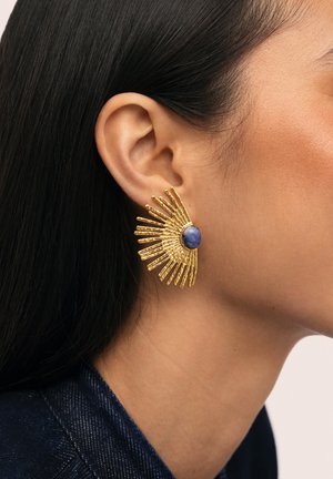 Pendiente dorado en forma de abanico con una piedra azul central, llevado en la oreja izquierda de una persona con cabello largo y oscuro.