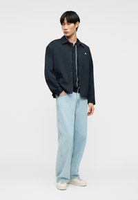 Νέος άνδρας που φοράει μαύρο σακάκι με κουμπιά πάνω από σκούρο πουκάμισο, ανοιχτό μπλε τζιν παντελόνι με φαρδιά μπάση και λευκά αθλητικά παπούτσια, στέκεται με το ένα χέρι στην τσέπη.