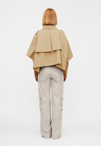 Poncho surdimensionné beige avec un design superposé et des pattes d'épaule, associé à un pantalon cargo de couleur claire avec des poches latérales.