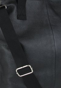 Zwarte lederen tas met een matte afwerking en een gestructureerd oppervlak. Voorzien van een zwarte stoffen riem en een metallic zilveren gesp. Eenvoudig en functioneel ontwerp.