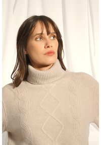 Hellbeige Rollkragenpullover mit geripptem Kragen und strukturiertem Strickmuster, das erhabene Zopfmuster aufweist. Weicher, voluminöser Stoff.