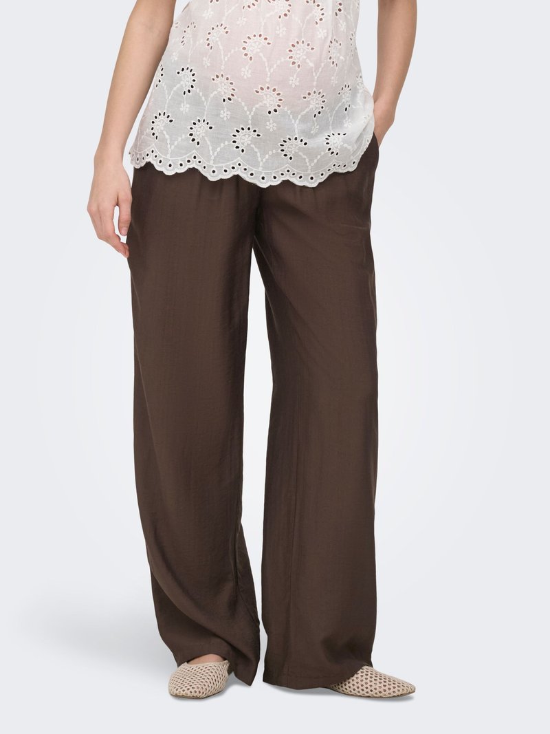 Femme portant un pantalon marron ample, un haut blanc brodé et des chaussures tissées beige à enfiler, debout avec une main dans la poche.