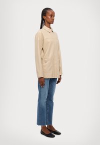 Lichtbeige jas met een kraag, twee voorzakken en een relaxte pasvorm, gecombineerd met blauwe high-waisted jeans en zwarte ballerina's.