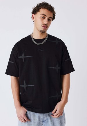 Giovane uomo con capelli ricci che indossa una t-shirt nera oversize con motivi a stella, una collana a catena d'argento e jeans di denim azzurro chiaro.