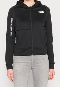 Sudadera con cremallera negra hecha de tejido suave con capucha. Presenta un logo en el pecho y texto a lo largo de la manga. Terminada con puños acanalados.