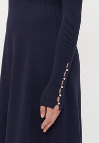 Abito a maglia ribbed blu navy caratterizzato da maniche lunghe con dettagli a bottone sui polsini e aperture vicino ai polsi. Texture liscia in tutta la sua lunghezza.