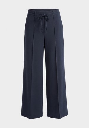 Pantalon ample bleu marine avec plis à l'avant et ceinture en tissu nouée à la taille sur un fond clair uni.
