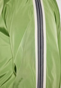 Giacca in satin verde con una finitura liscia, caratterizzata da una striscia bianca e blu scuro lungo la spalla e la manica.