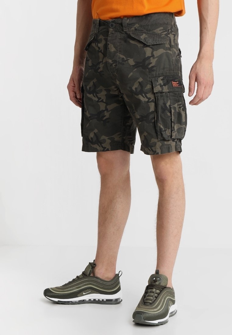 Pantaloni cargo camouflage in verde scuro e nero con molte tasche, tessuto resistente e una vestibilità comoda. Indossati con sneakers verde oliva.