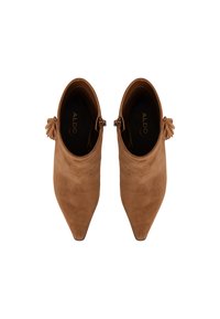 Botas de tobillo de ante en tono beige con puntera afilada, que cuentan con un borlón decorativo en el lateral y una textura suave. Logo de la marca visible en el interior.