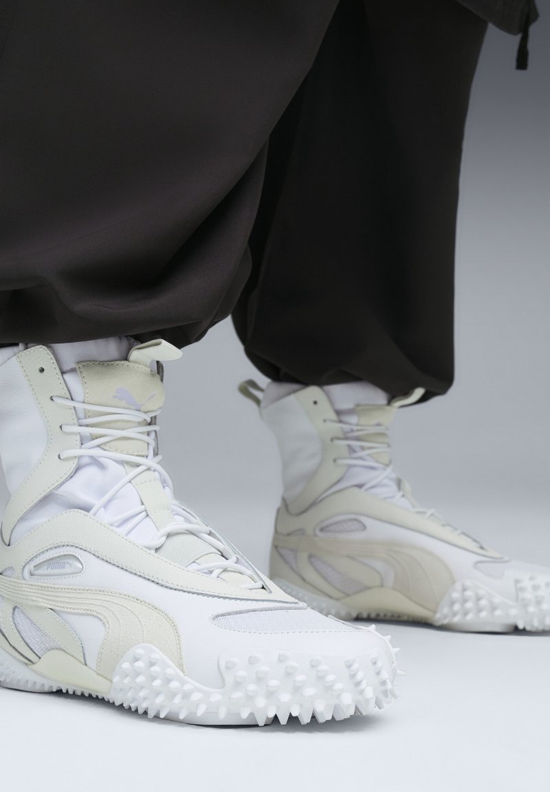 Baskets montantes blanches et beiges avec des semelles à pointes texturées, portées avec un pantalon noir ample sur un fond gris clair.