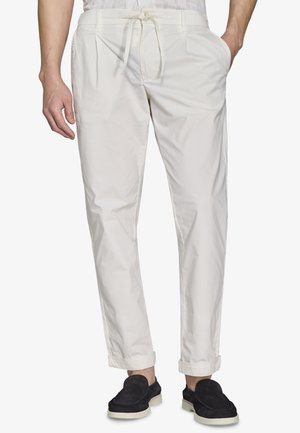 Męskie kremowe spodnie ze ściągaczem, z przodu z zakładkami, mankietami u dołu, kieszeniami po bokach, w zestawieniu z czarnymi wsuwanymi butami i białą koszulą.