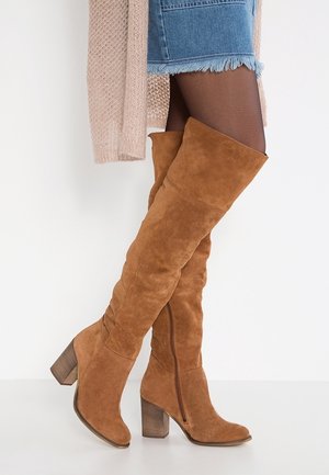 Bruine suède laarzen over de knie met blokhakken, gedragen met zwarte panty's en een gerafelde denim rok, gecombineerd met een beige gebreid vest.