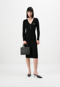 Woman in black long-sleeve midi dress with gold buttons holding black studded handbag, wearing black pointed-toe kitten heels on white background.