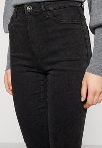 Jeans negros de tiro alto, hechos de denim, con bolsillos delanteros, una textura suave y un ligero acabado deslavado. Herrajes de botón plateado visibles.