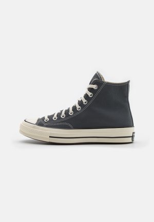 Schwarzer High-Top-Leinenschuh mit weißen Schnürsenkeln, weißer Gummisohle und schwarzem Streifendetail, im linken Seitenprofil auf weißem Hintergrund gezeigt.