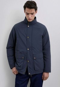 Tummansininen toppatakki, jossa on korkea kaulus, etuvetoketju ja napitus, sekä kaksi etutaskua ja kullanvärisiä yksityiskohtia.