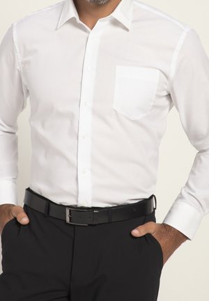 Mand iført en hvid skjorte med lange ærmer og knapper, med sort bælte og sorte bukser, hænderne i lommerne.