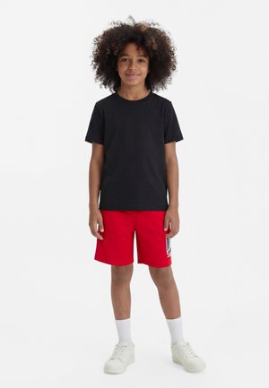 A gyermek előre néz állva, fekete pólót, piros rövidnadrágot fehér logóval, fehér zoknit és fehér tornacipőt visel fehér háttéren.