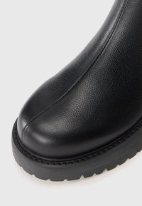 Gros plan d'une botte en cuir noire montrant un bout arrondi, des coutures visibles et une semelle en caoutchouc épaisse avec un motif de relief sur fond blanc.