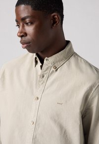 Lichtbeige button-up overhemd met een kraag, voorzien van een subtiele merklogo en standaard knoopsluiting. Zacht, gestructureerd materiaal.