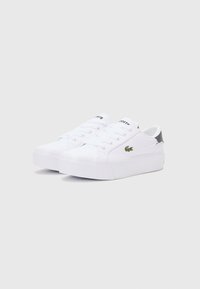 Balti odiniai sportiniai bateliai su tekstūriniu guminio pado, demonstruojantys raištelių uždarymą ir žaliu Lacoste logotipu šone. Papuošti pilkos spalvos detalėmis.