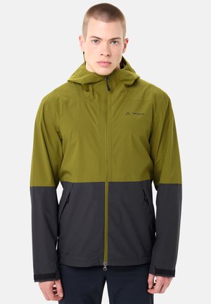 Junger Mann mit kurzem Haar, der eine zweifarbige olivgrüne und dunkelgraue Jacke mit Reißverschluss und Kapuze trägt und vor einem einfarbigen Hintergrund steht.