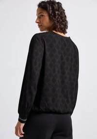Femme aux cheveux bouclés portant un blouse noire à manches longues avec un motif géométrique subtil et un pantalon noir ajusté, tournant légèrement sur le côté.