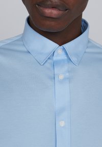 Chemise bleu clair à boutons avec un tissu texturé, col pointu et boutons blancs, visibles au niveau du cou. Design simple et classique.