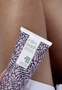 Il tubo del detergente intimo presenta un design rosa a motivi con foglie scure, testo bianco sull'etichetta ed è posizionato accanto a una superficie della pelle liscia.