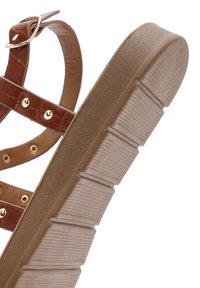 Brown synthetic sandals with gold-tone accents, featuring a textured sole, adjustable strap, and multiple crisscrossing decorative straps.