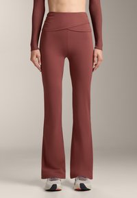 Donna che indossa pantaloni da yoga a zampa di elefante color ruggine a vita alta con dettaglio di fascia incrociata e scarpe da ginnastica bianche su sfondo semplice.