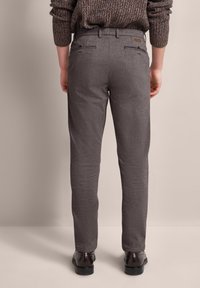 Graue Fischgrat-Hosen mit geradem Schnitt, ausgestattet mit einem Bund, Gürtelschlaufen und zwei Gesäßtaschen. Getragen mit braunen Schuhen vor einem neutralen Hintergrund.