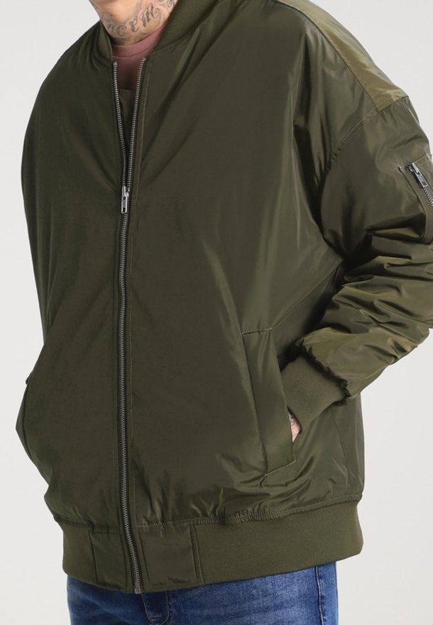 Homme portant une veste bomber vert olive avec fermeture éclair sur le devant, poches latérales et poche zippée sur la manche, associée à un jean bleu.