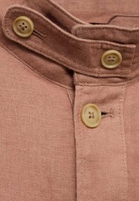 Gros plan d’un col de chemise marron avec deux boutons beige sur le col et un bouton beige sur le devant de la chemise.
