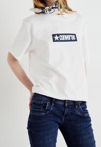 T-shirt en coton blanc avec un graphique rectangulaire bleu portant le mot "CONVERSE" et une étoile. Assorti à un jean en denim bleu foncé.
