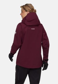 Vinröd vattentät jacka med huva, dragkedjeförsedda fickor och justerbara ärmslut. Har logga på ryggen och slät yta. Svarta handskar används.