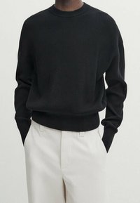 Knitted black sweater with a crew neckline, ribbed texture, and loose fit, paired with light-colored tailored trousers.