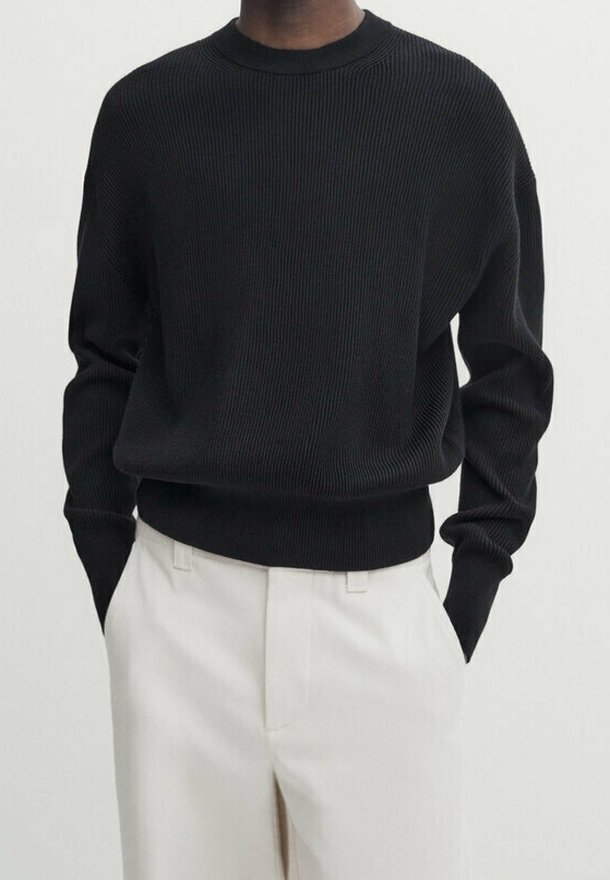 Knitted black sweater with a crew neckline, ribbed texture, and loose fit, paired with light-colored tailored trousers.