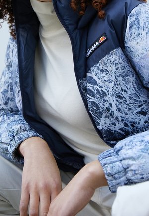 Osoba ubrana w niebiesko-białą, marmurkową kurtkę z elastycznymi mankietami, granatowym kołnierzem i naszywką z logo „ellesse”, w połączeniu z beżową bluzką i spodniami.