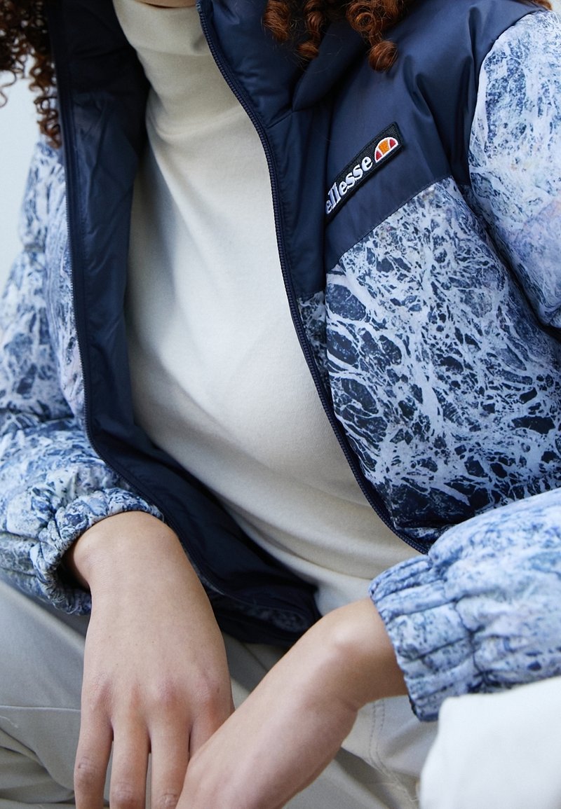 Personne portant une veste marbrée bleu et blanc avec des poignets élastiques, un col bleu marine et un écusson avec le logo "ellesse", assortie d'un haut beige et d'un pantalon beige.