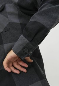Zwart en grijs geruit overhemd met een knoopmanchet, subtiele textuur en een patchzak op de voorkant. De stof lijkt zacht en duurzaam te zijn.