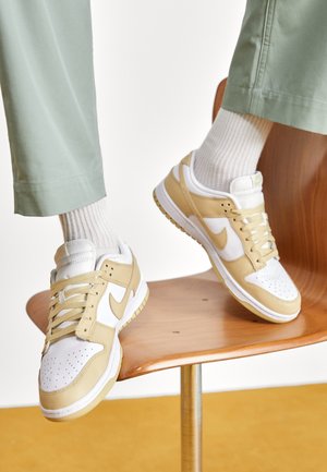 Osoba nosząca beżowo-białe buty sportowe Nike z białymi skarpetkami i jasnoliczonymi zielonymi spodniami, siedząca na drewnianym krześle z uniesionymi nogami.