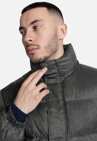 Sötétszürke párnázott dzseki, magas gallérral és texturált felülettel. A mandzsetták bordás sötétkék színűek. A modell finoman megérinti a gallért.