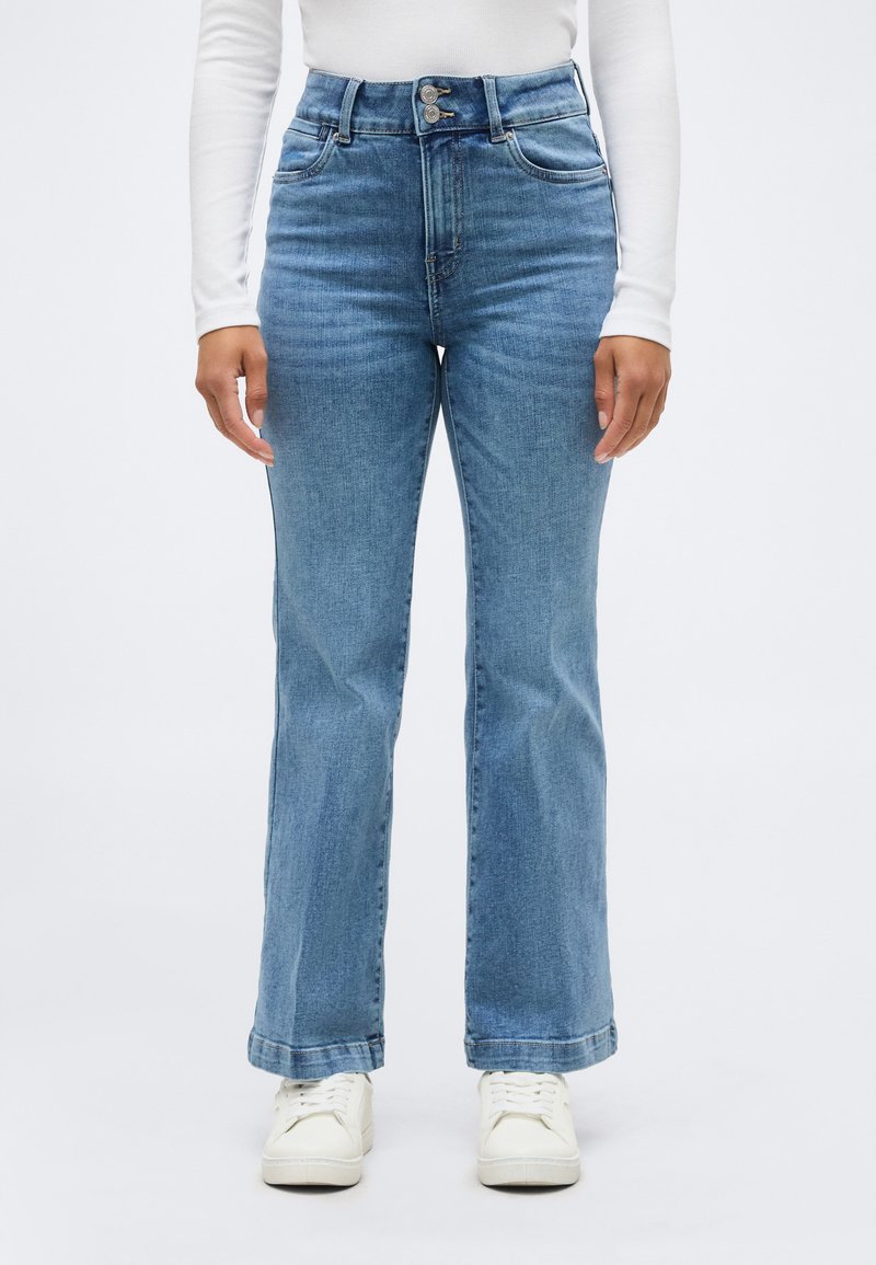 Άτομο που φοράει τζιν ψηλόμεσο ανοιχτού μπλε χρώματος με διπλό κούμπωμα και λευκά αθλητικά παπούτσια, στέκεται μπροστά σε απλό φόντο.