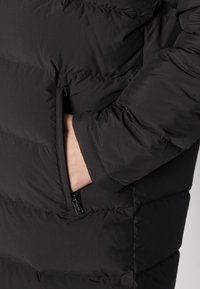 Chaqueta de plumas negra con textura acolchada, bolsillo lateral con cremallera y puños elásticos. El material parece suave e insulated.
