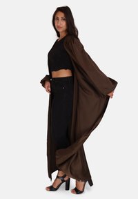 Bruine satijnen lange jas met brede mouwen, gedragen over een zwart cropped top en hoge taille zwarte spijkerbroek, gecombineerd met zwarte hakken.