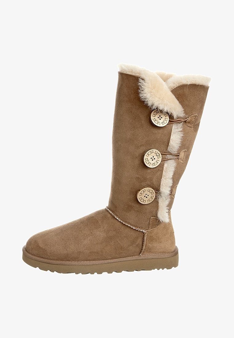 Zamszowy beżowy botek z trzema ozdobnymi guzikami, wyściółką z owczej wełny i płaską gumową podeszwą. Posiada wysokość do połowy łydki oraz przeszywane detale.
